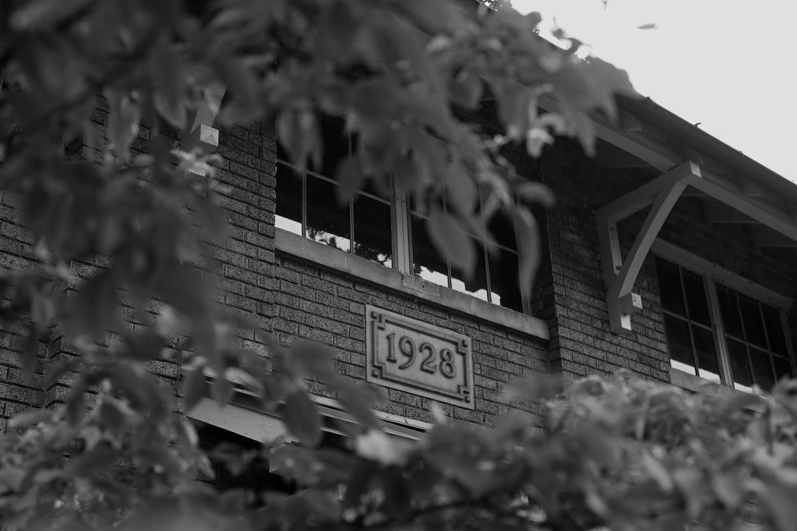 Grayscale image of the front of a brick building with the year '1928' seen through foliage